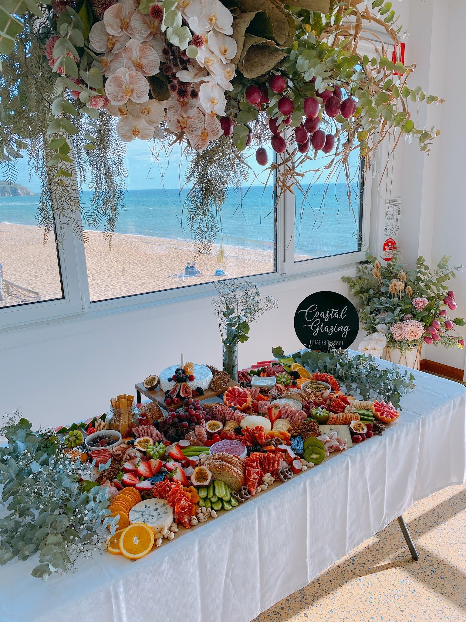 Coastal Grazing table covered in charcuterie and fruit, with beach and ocean views through windows behind it